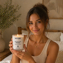 Woman holding a candle labeled 'Enchanted Forest' in a cozy indoor setting.