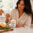 Woman lighting a Fizzy Citrus candle in a kitchen with oranges and mint on a cutting board.