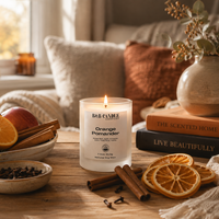 A lit B&B Candle 'Orange Pomander' in a frosted glass jar sits on a wooden table surrounded by orange slices, cinnamon sticks, dried orange slices, and books in a cozy, warm-toned living room setting.