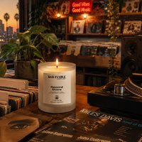 A lit white candle labeled 'BAB CANDLE Record Store' sits on a wooden table surrounded by vinyl records, a turntable, plants, and a record store interior with a city skyline at sunset in the background.
