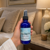 Hand holding a blue spray bottle labeled B&B Candle Co. Rest & Relax and is in a bedroom setting.