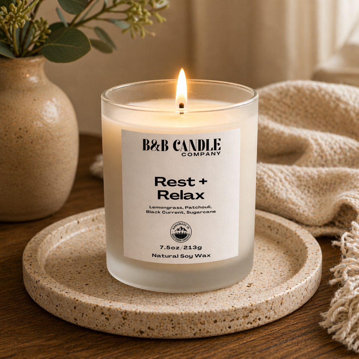 Candle labeled 'B&B Candle Company Rest + Relax' on a textured surface with a vase and blanket in the background.